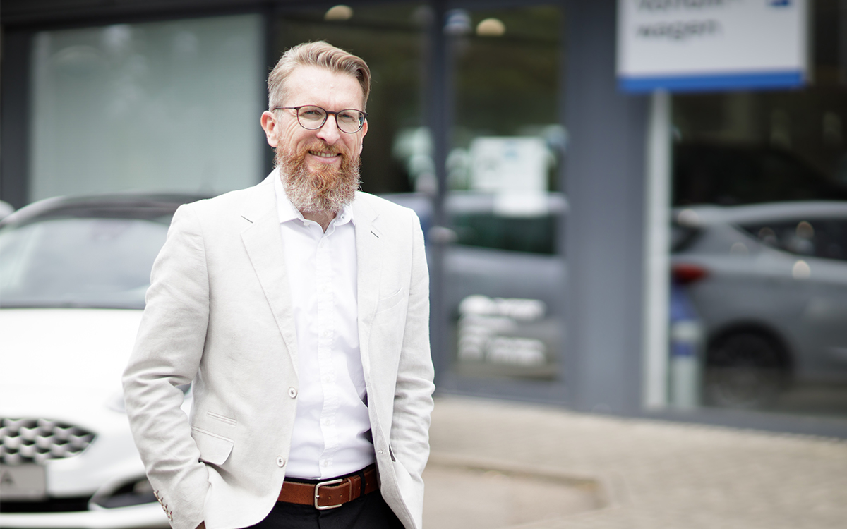 Autoverkaeufer Juergen Speth vom Autohaus Zwerger draussen mit hellem Sakko, weissem Hemd braunem Guertel und dunkler Hose